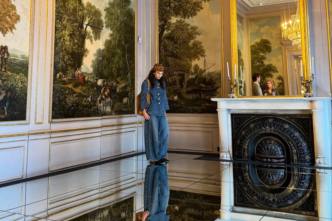 A person in denim clothing stands in an ornate room with floor-to-ceiling murals and reflective flooring.