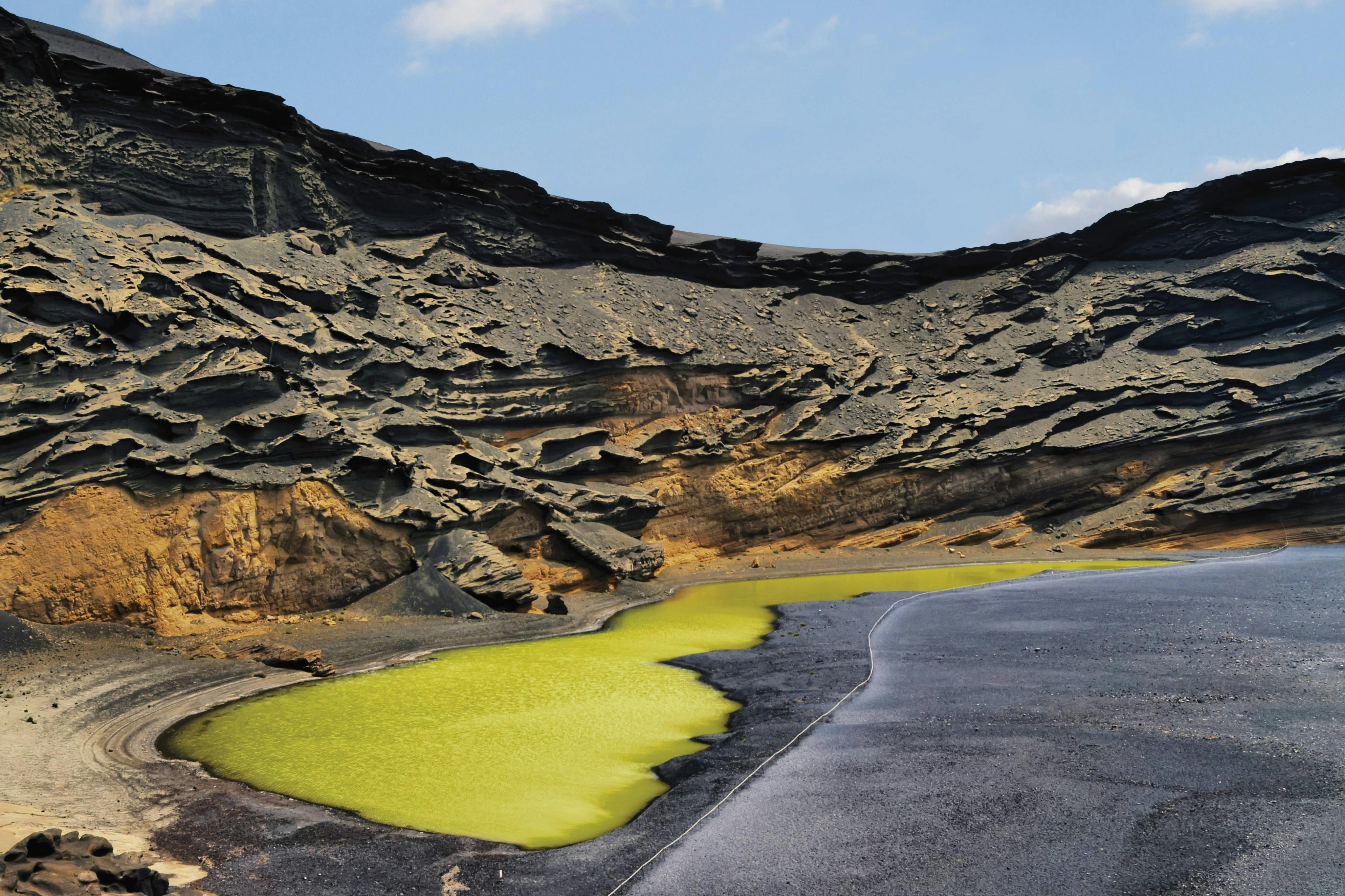 Jagged black and brown rocks surround a small, bright green sulfuric lake under a clear blue sky.