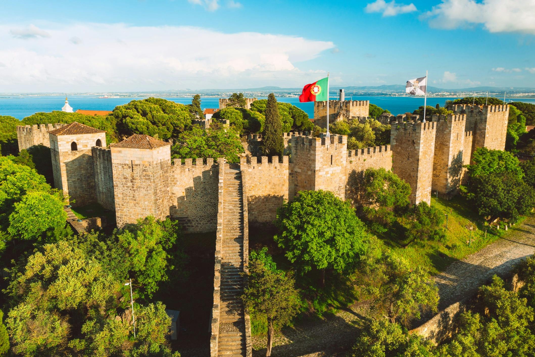 A historic castle with stone walls, trees, and flags, set against a backdrop of blue sky and distant water.