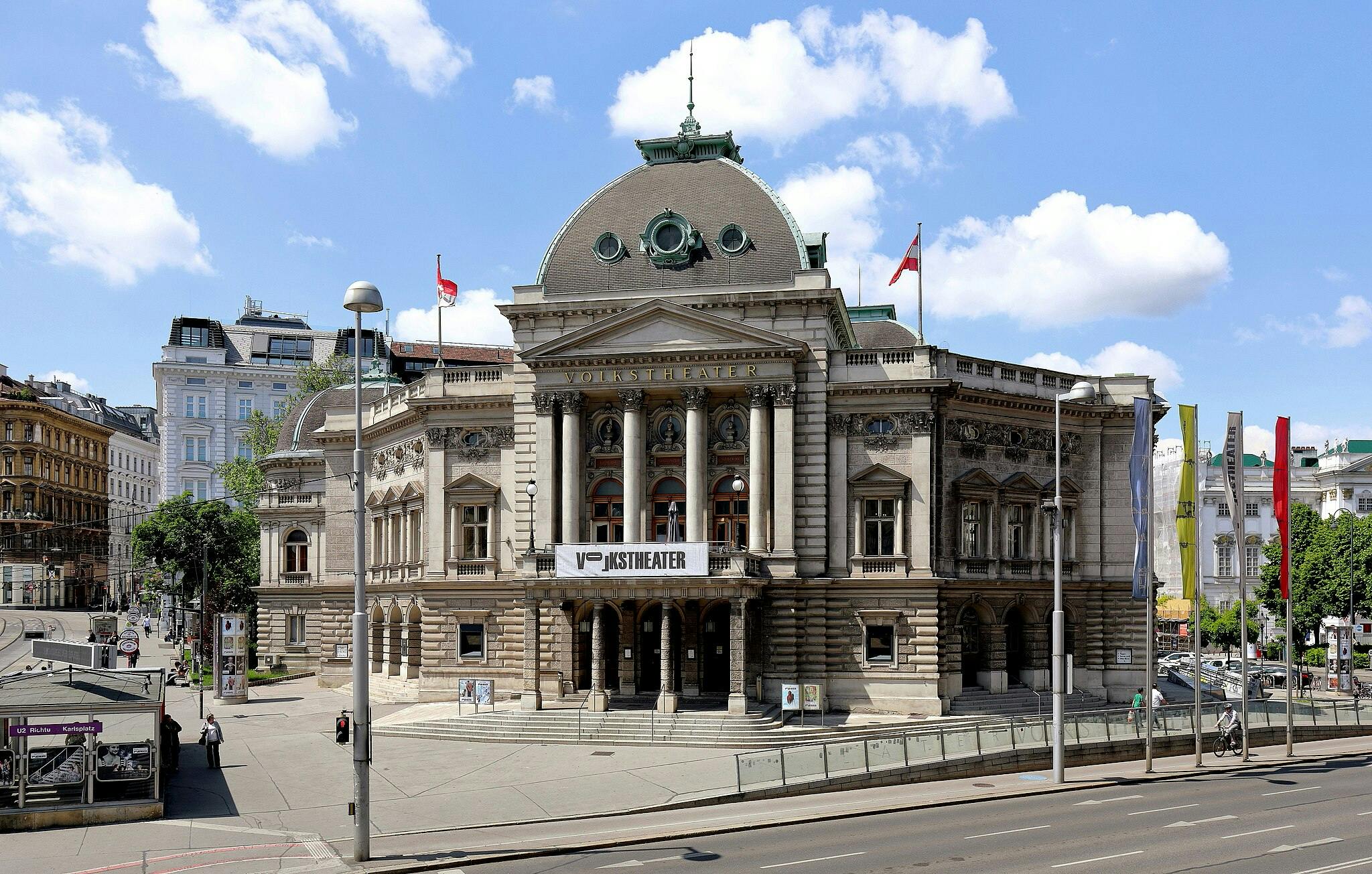 Historic building with classical architecture, labeled "Volkstheater," adorned with flags, columns, nearby street, and pedestrians.