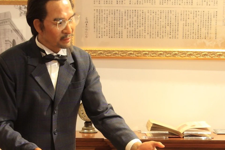 A museum exhibit featuring three life-size wax figures seated around a wooden table, with framed photos and documents on the wall.