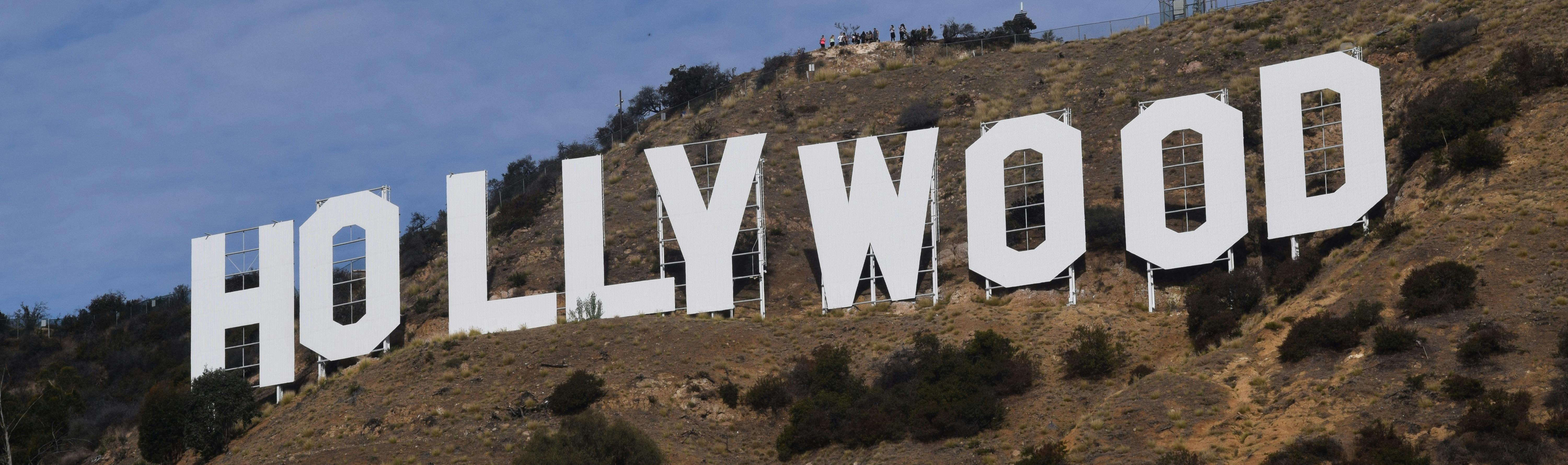 这座标志性的好莱坞标志位于山坡上，蓝色略微多云的天空下，背景中可以看到几个人。