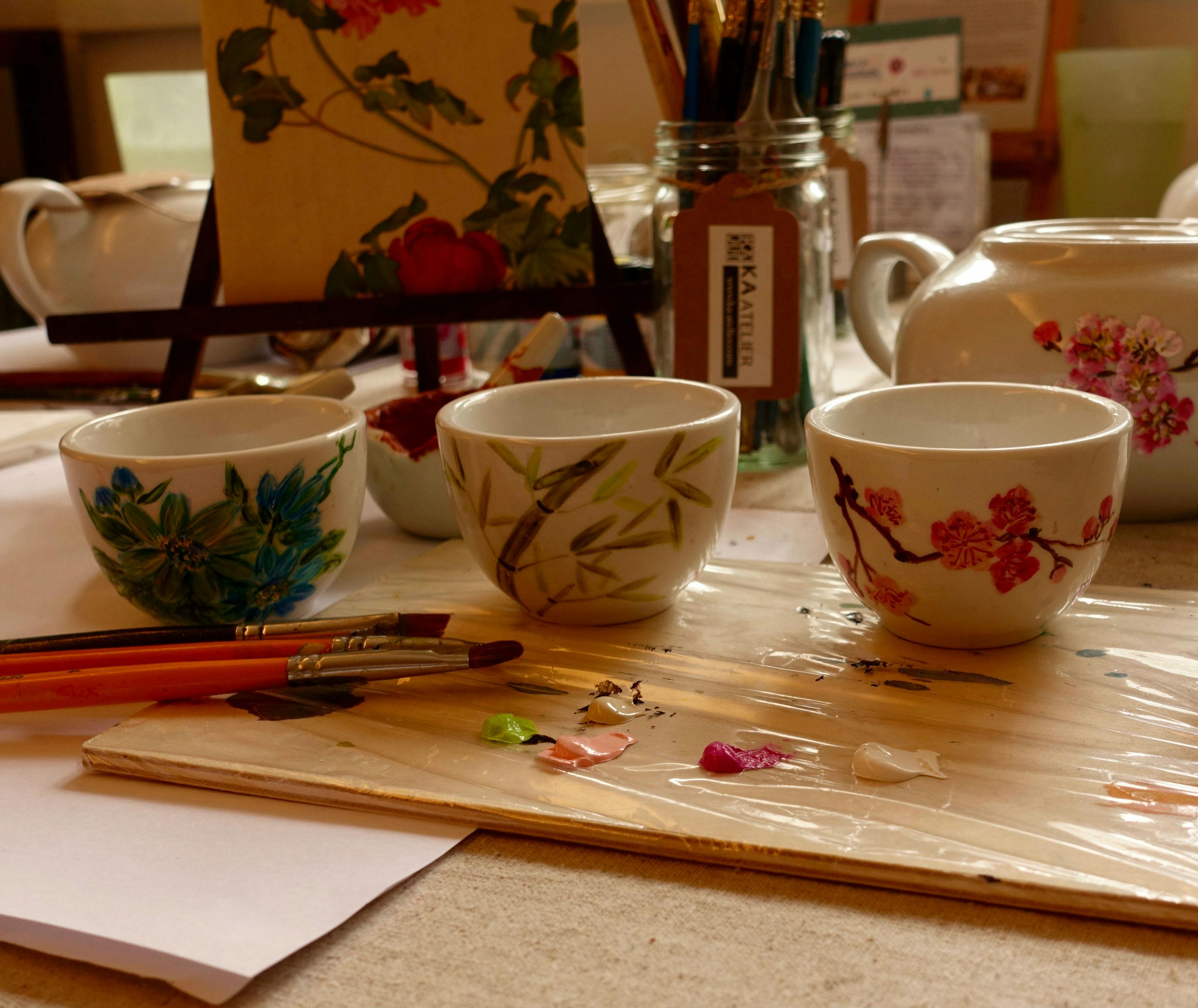 三只彩绘茶杯，配有花卉图案、画笔、调色板、罐子和桌面上的茶壶，用于绘画。