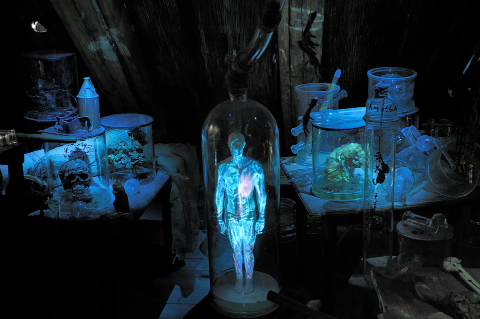 A glowing holographic figure in a glass cylinder, surrounded by various scientific equipment and containers in a dimly lit room.