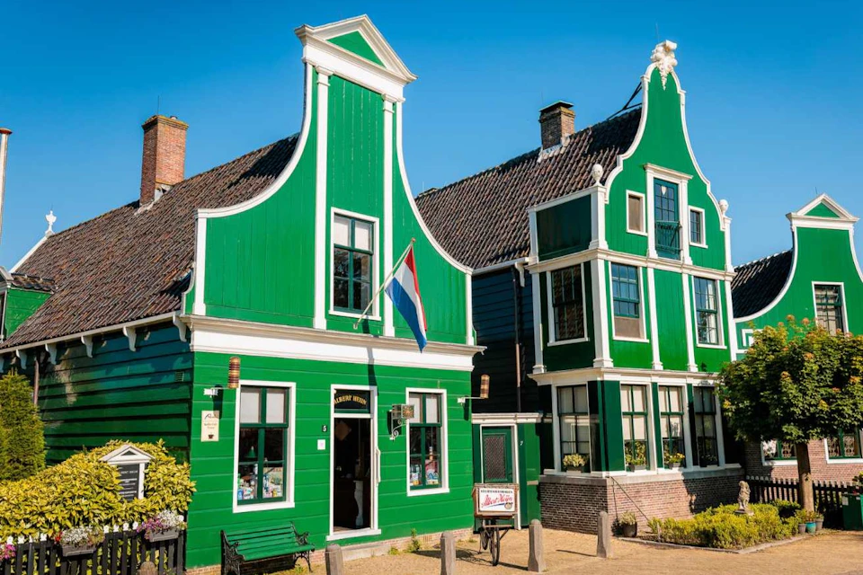 Two traditional green Dutch houses with stepped gables, a flag hanging between them, under a clear blue sky.