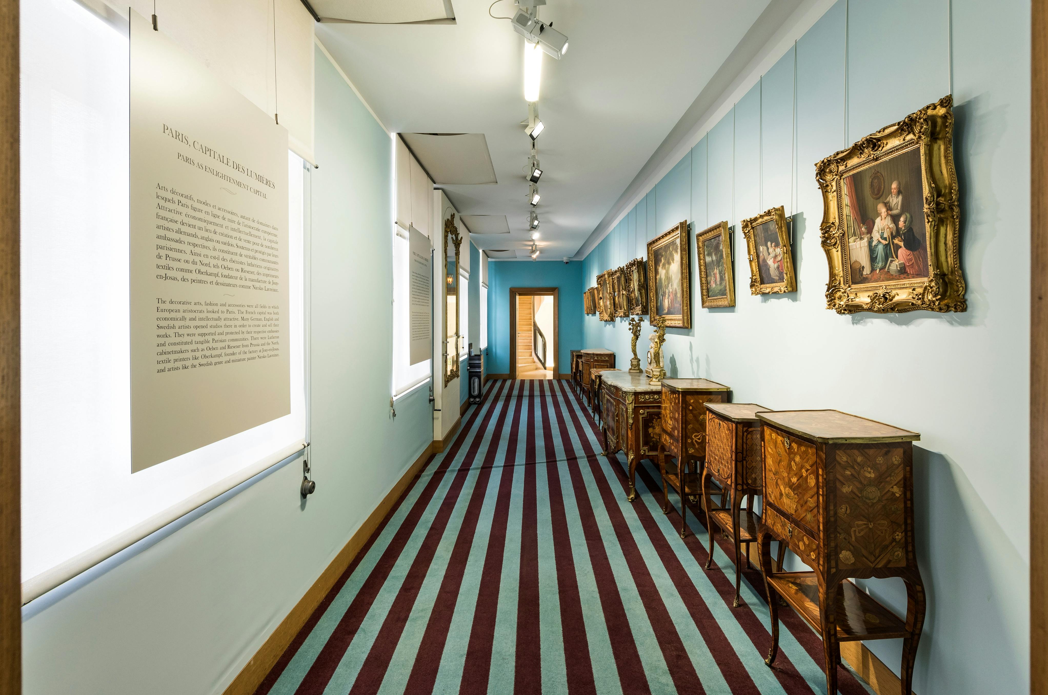 博物馆走廊设有框架画，右墙设有华丽的桌子，尽头设有条纹地毯，通往木门。