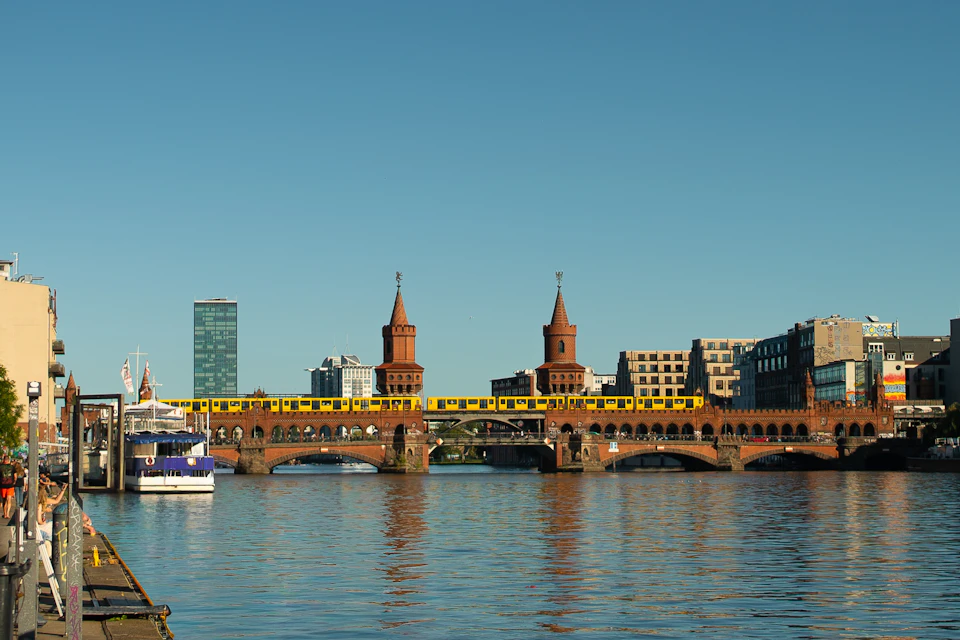 A yellow train crosses the Oberbaum Bridge over a river, with city buildings in the background under a clear blue sky.