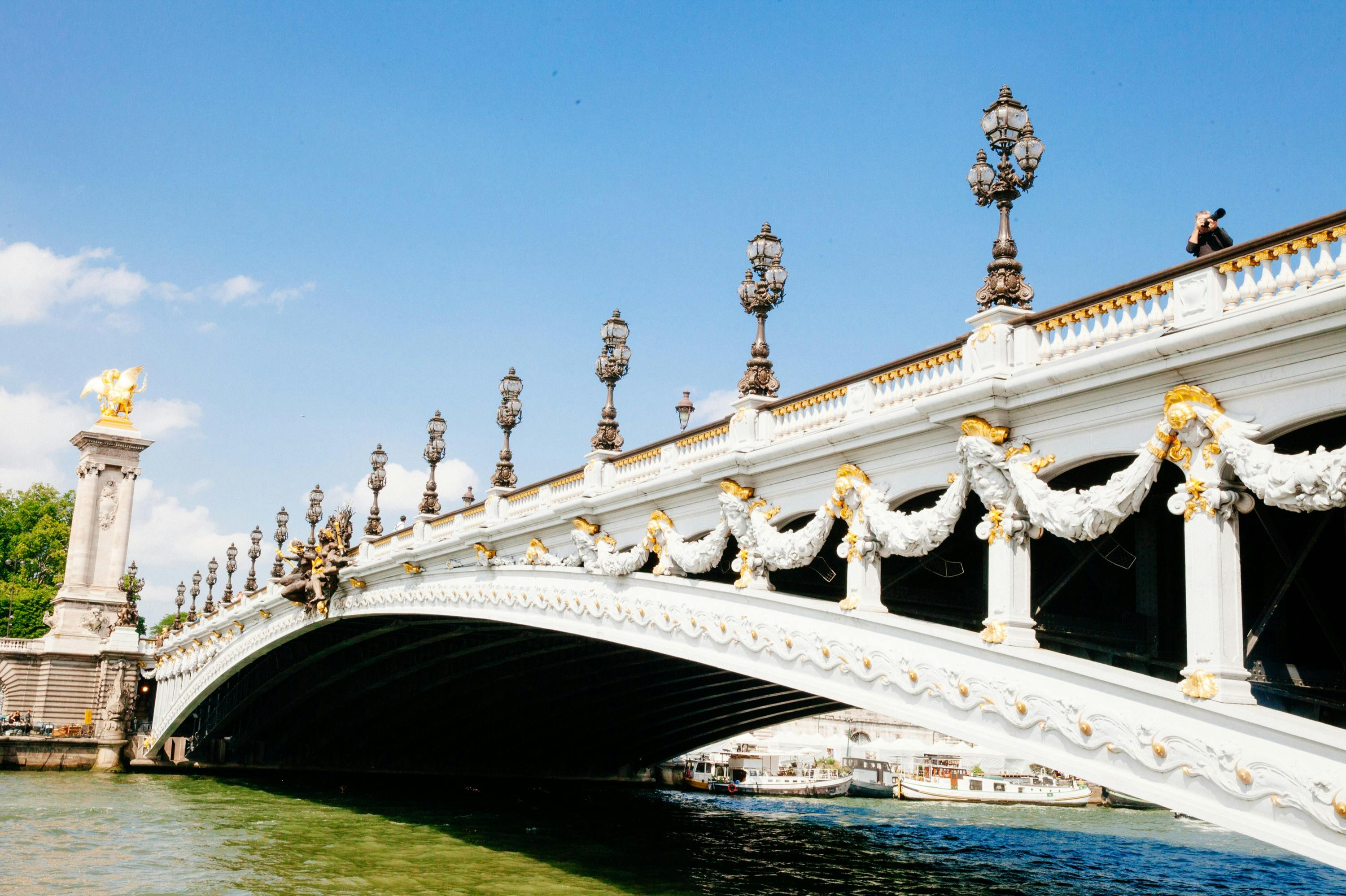Pont orné de blanc et d'or avec des lampadaires décoratifs, des statues et une personne en train de photographier, le tout sur fond de ciel bleu.