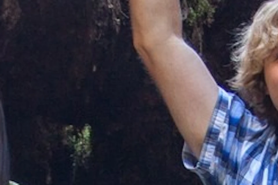 Two people stand in front of a large, uprooted tree stump in a dense forest, both smiling at the camera.
