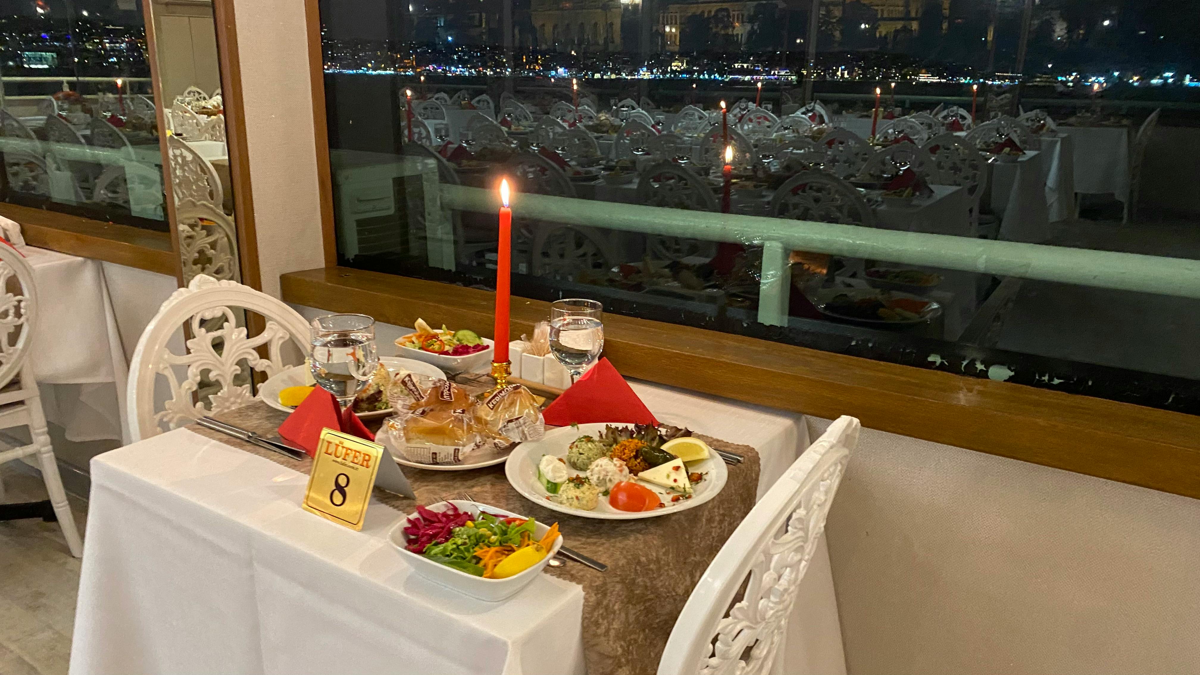 A restaurant table set for two with plates of food, a red candle, and a view of a cityscape across a waterfront at night.