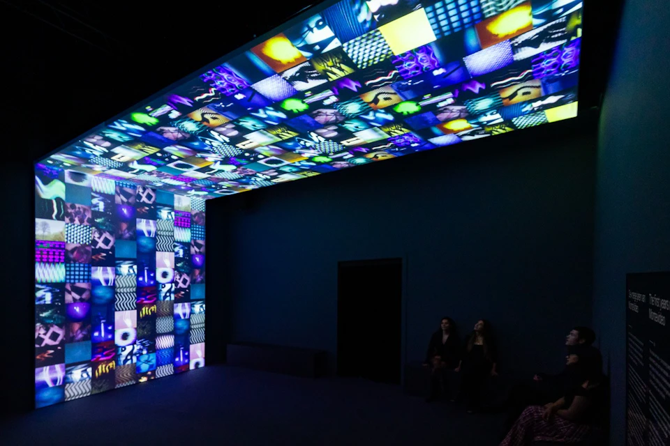 A dark room with colorful, patterned light projections on the ceiling and walls, and two people sitting on a bench.