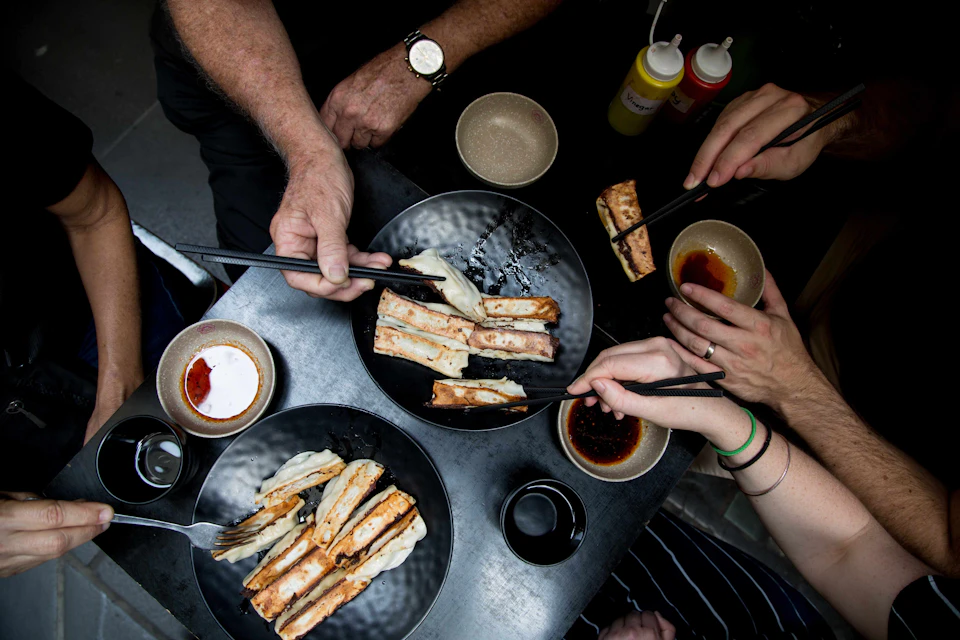 Three people using chopsticks to eat gyoza from black plates on a dark table, with sauces and condiments around.