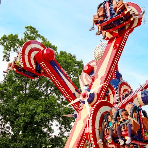 People enjoying a colorful amusement park ride with spinning seats, against a backdrop of trees and a blue sky.