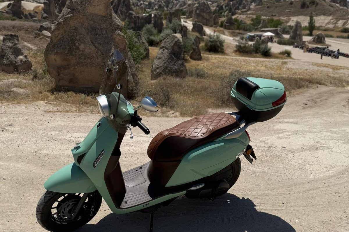 一辆薄荷绿色踏板车，配有棕色绗缝座椅和后部储藏室，停在尘土飞扬的小路上，背景是岩石地形。