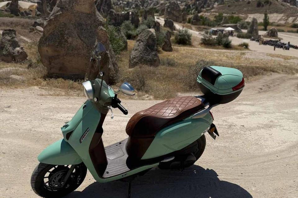 A mint-green scooter with a brown quilted seat and rear storage parked on a dusty path with rocky terrain in the background.