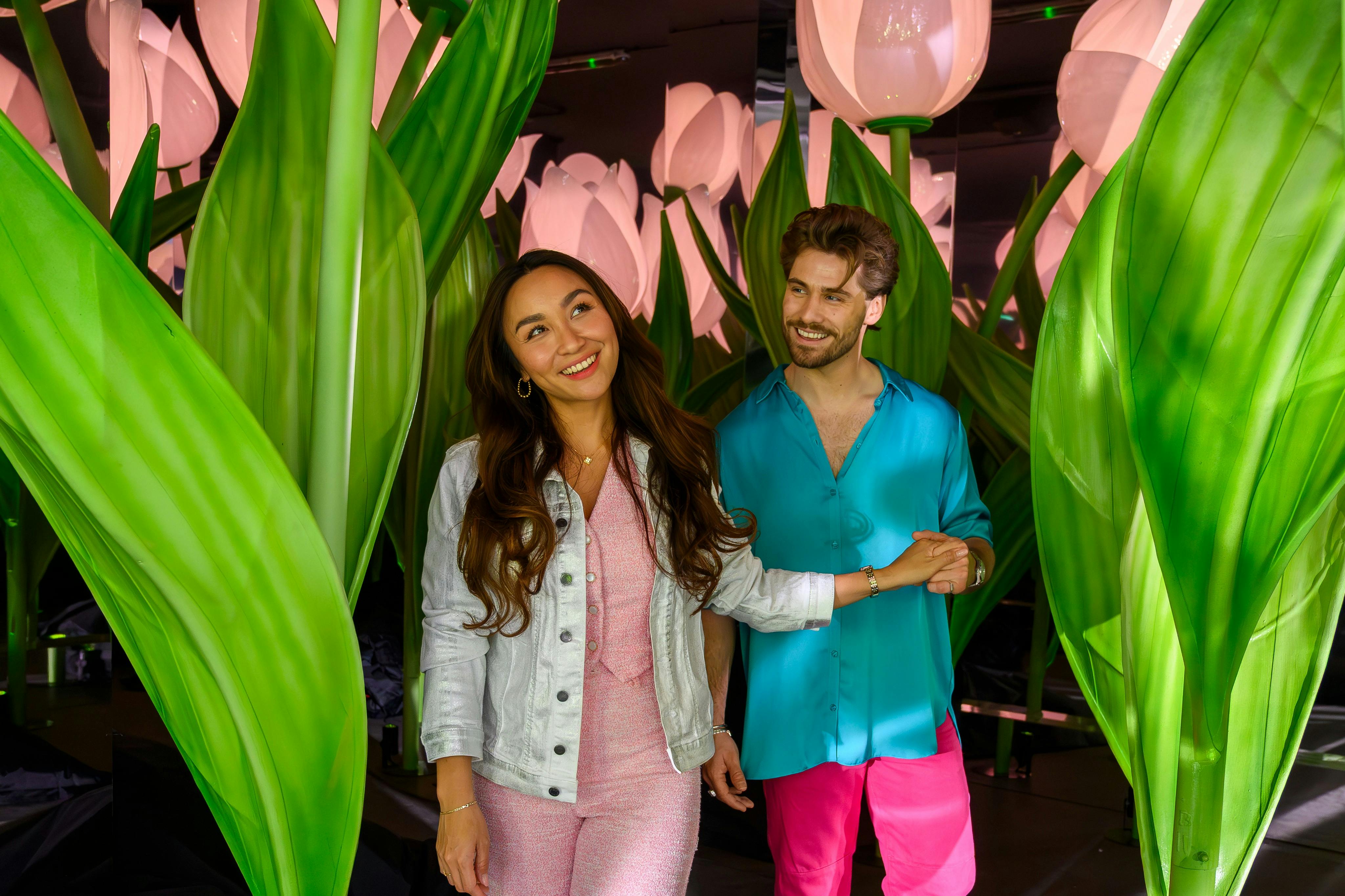Ett leende par som går genom en uppvisning av stora, upplysta tulpaner och gröna löv i en inomhusmiljö.