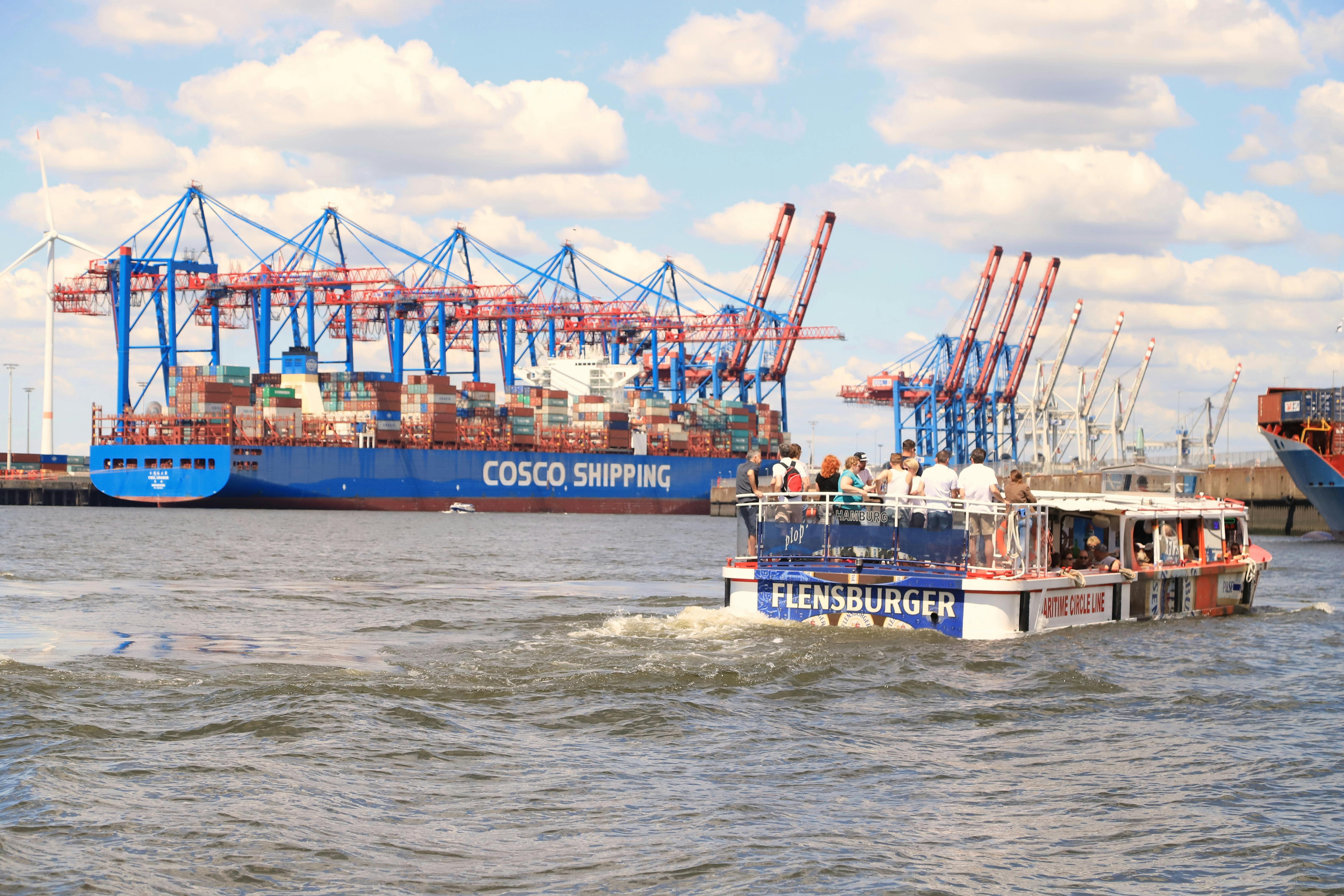 Un bateau nommé "FLENSBURGER" avec des personnes à bord navigue près de grands conteneurs d'expédition COSCO et de grues dans un port très fréquenté.