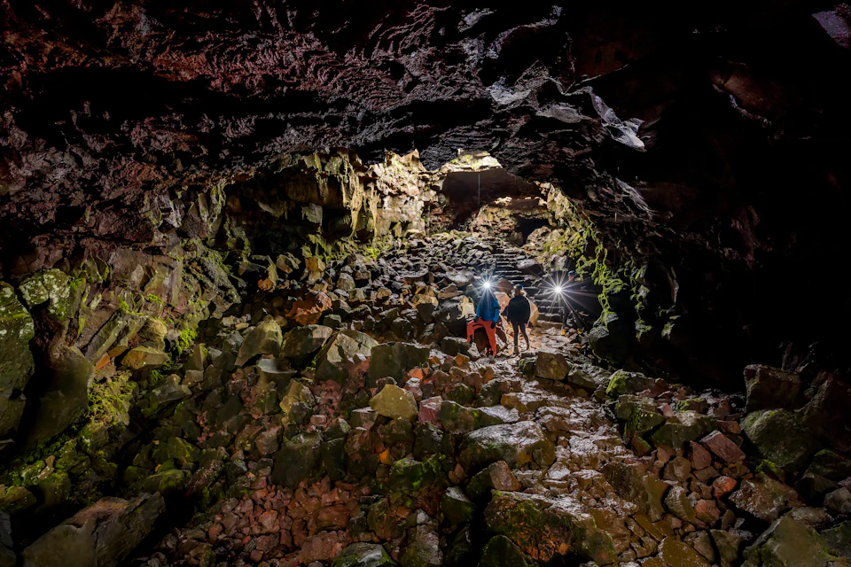 Four people exploring a rocky cave, illuminated by headlamps. The cave walls are rugged and covered in moss.