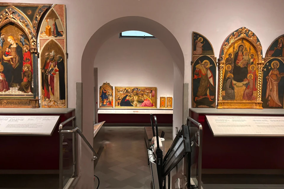 A museum gallery with religious artwork displayed on walls and a central archway leading to another exhibit space.