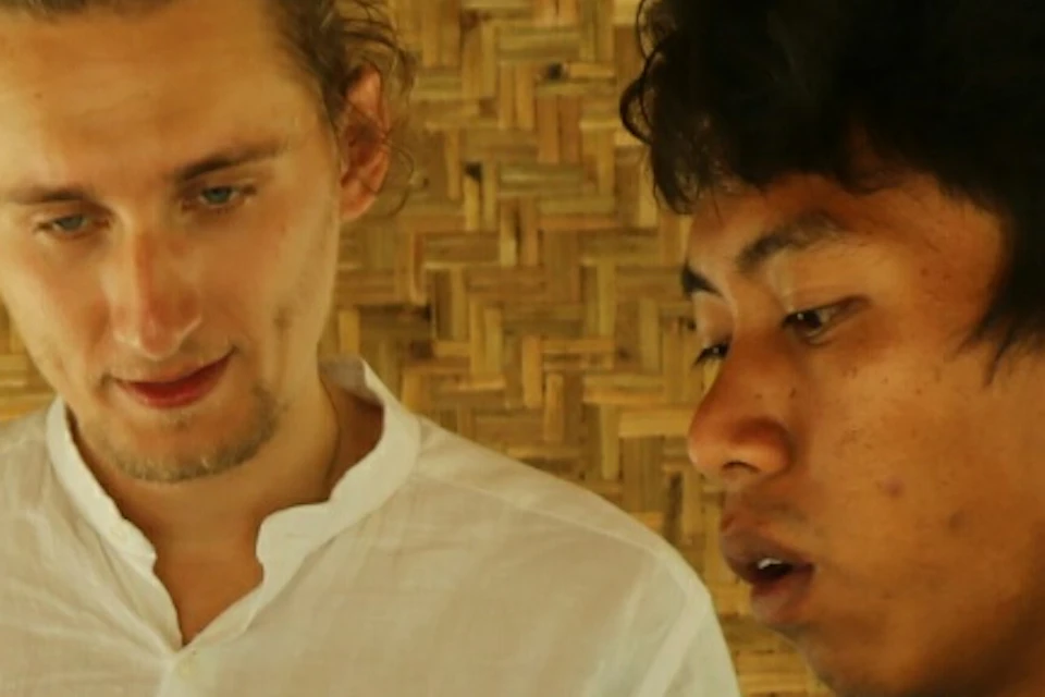 Three people examine dried cocoa beans on large trays in rustic, bamboo-walled setting; one in a white shirt, the other in a teal polo.