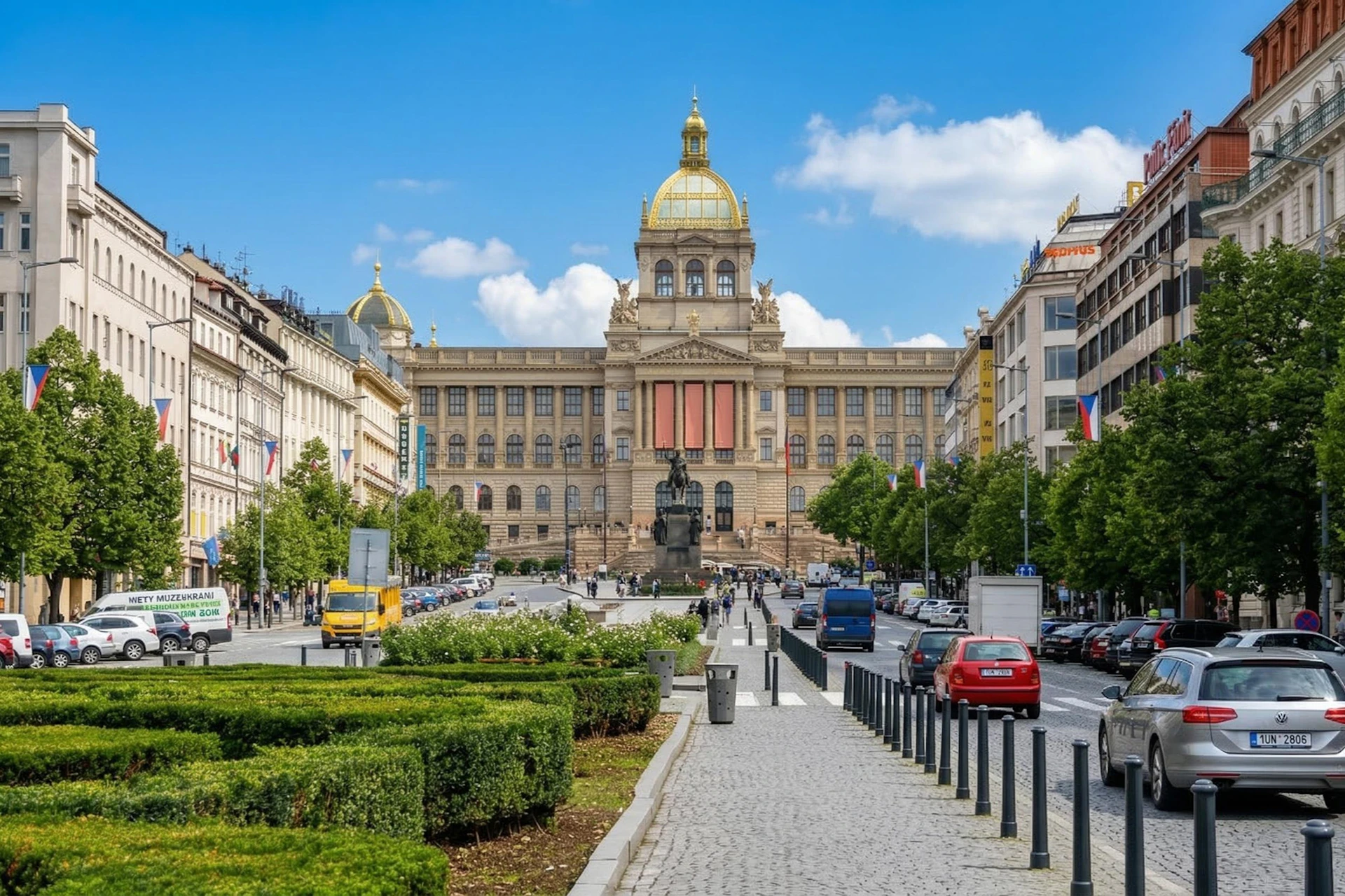A wide street with a pedestrian walkway leading to a large historic building with a golden dome, surrounded by trees and cars.