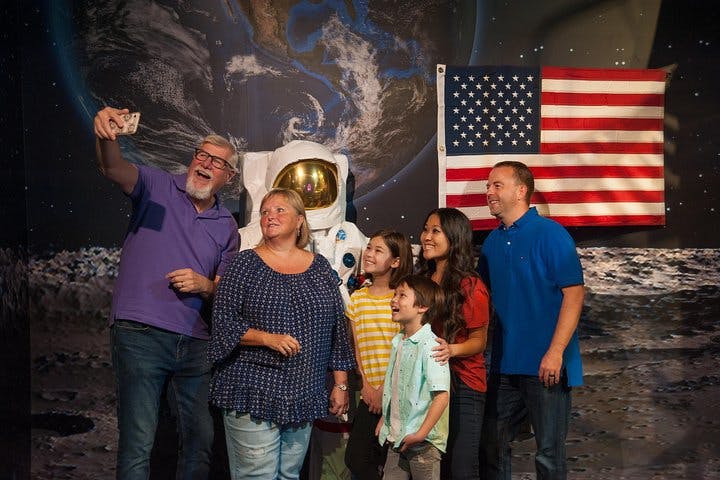 Skupina šesti lidí pózuje pro selfie s astronautským oblekem a americkou vlajkou v pozadí.