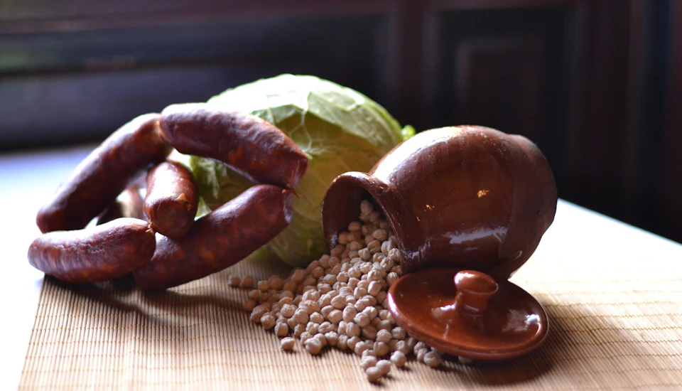 A clay pot spills beans next to a green cabbage and a ring of sausages on a woven mat.