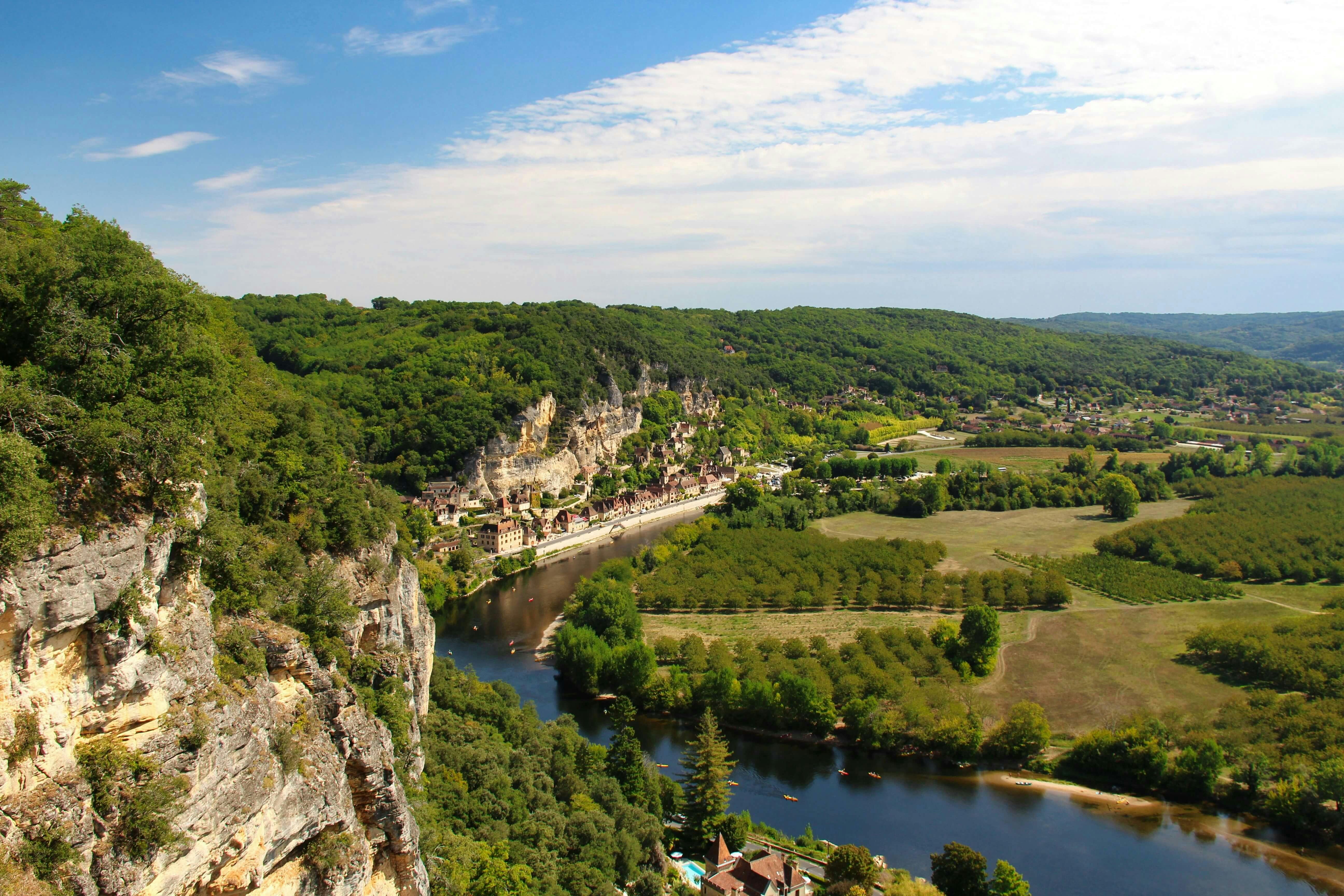 A river flows through a valley flanked by lush green hills and cliffs. A village sits at the base of the cliffs near the river.