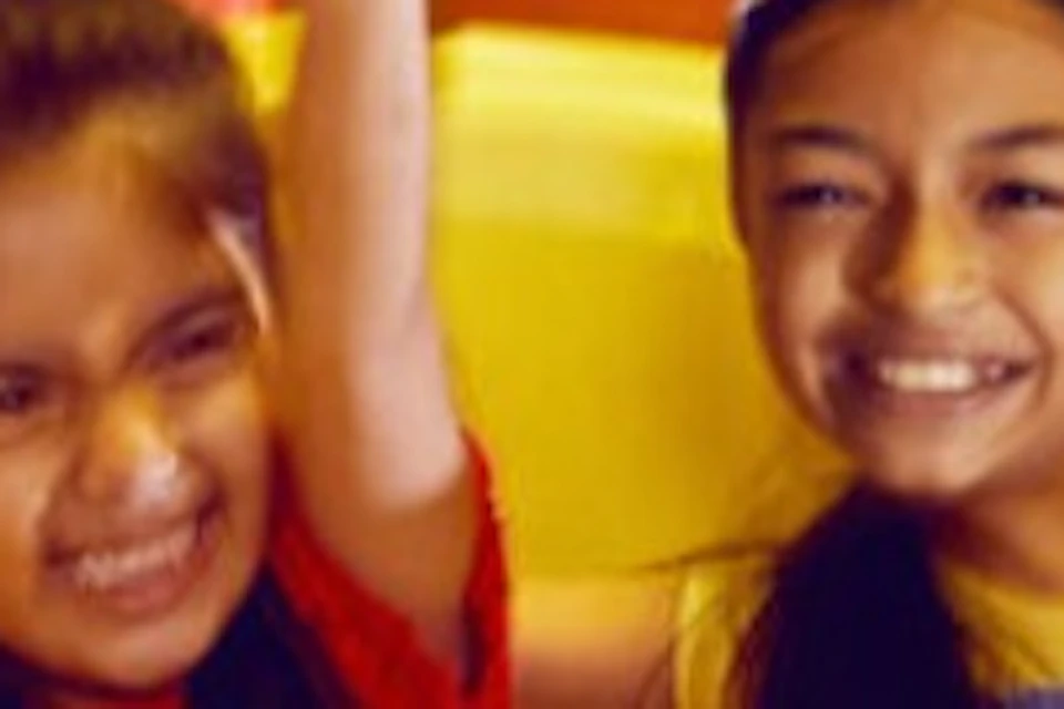 Two children smiling and raising their arms while riding colorful spaceship-themed amusement ride indoors.