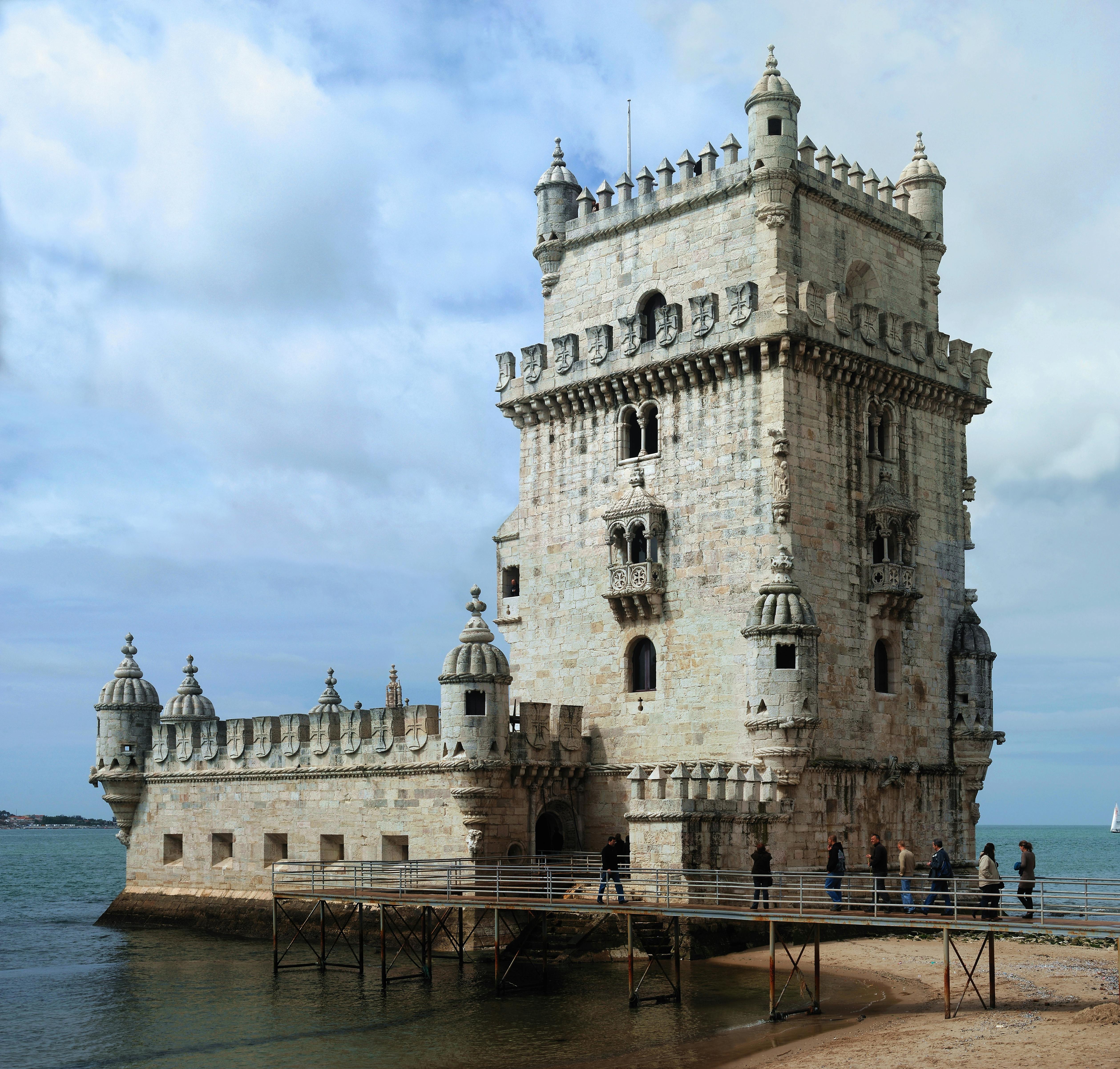 Stone tower with intricate details stands on the water's edge, connected to the shore by a wooden bridge. People walk along the bridge.