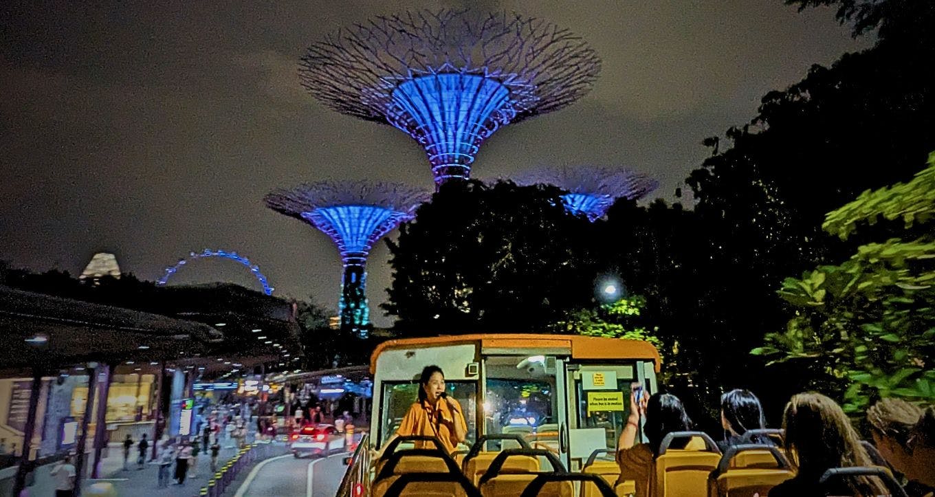 Turisté v otevřeném autobusu v noci s osvětlenými strukturami Supertree Grove v pozadí v Gardens by the Bay v Singapuru.