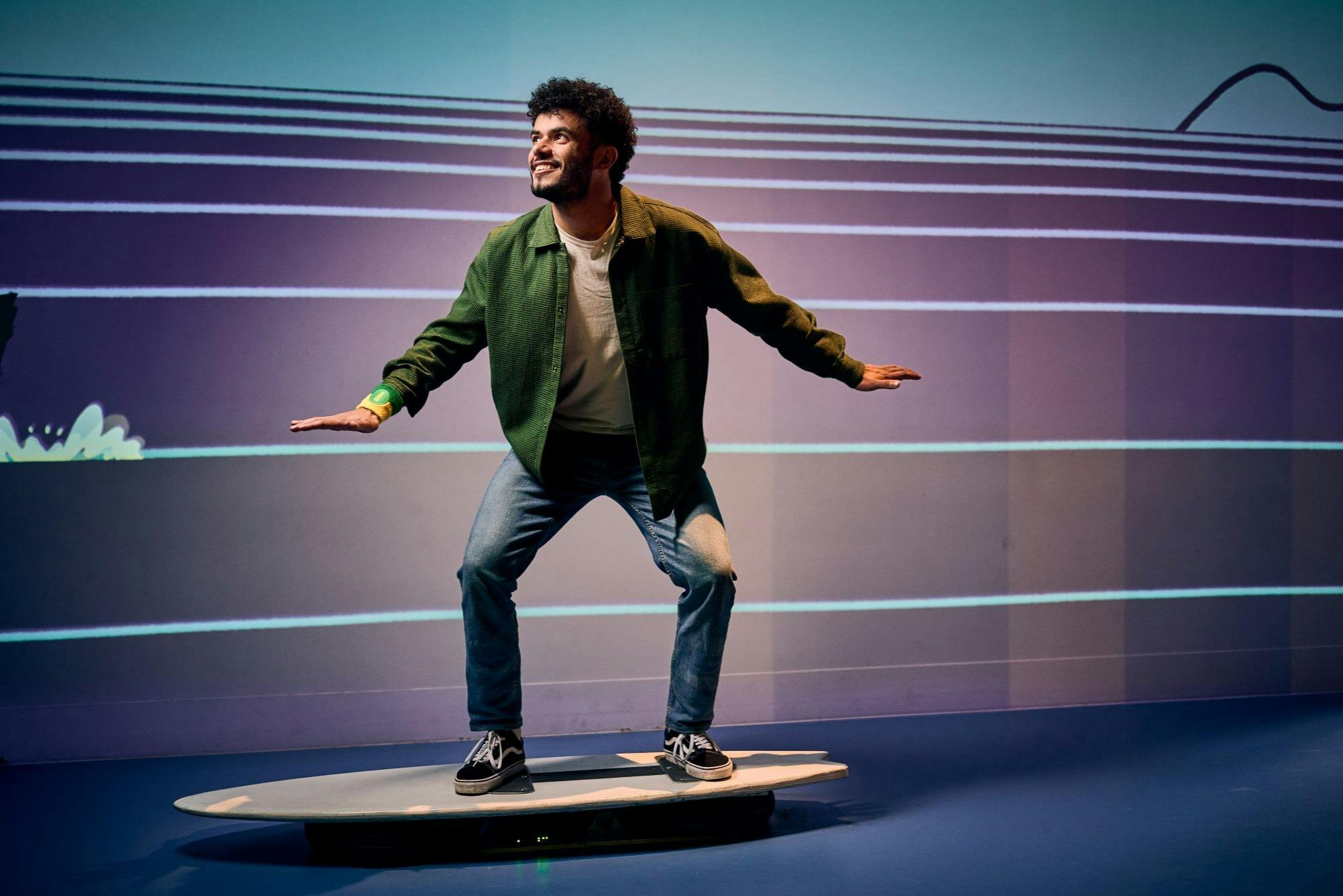 Une personne pose sur une plate-forme en forme de planche de surf à l'intérieur, simulant le surf, avec une toile de fond rayée et une tenue décontractée.