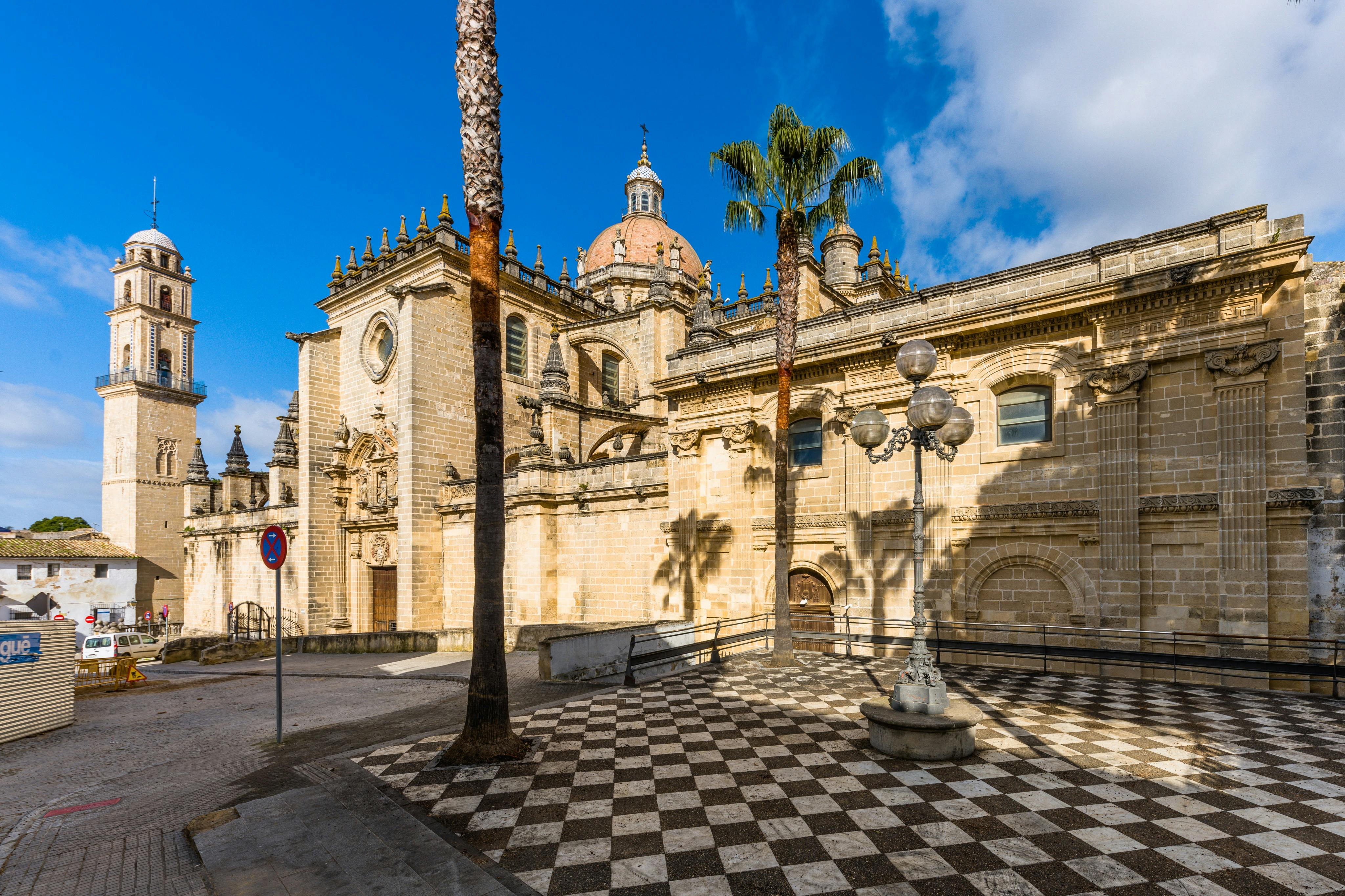 Bâtiment historique en pierre à l'architecture et aux dômes ornés, palmiers, place en damier et ciel d'un bleu éclatant.
