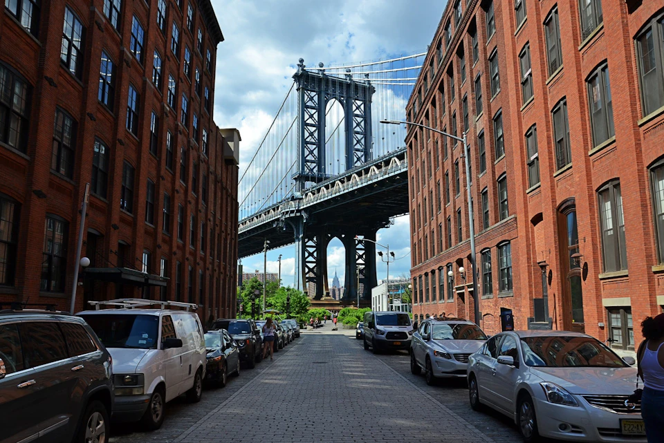 A street view framed by tall red brick buildings with the Manhattan Bridge in the background under a partly cloudy sky.
