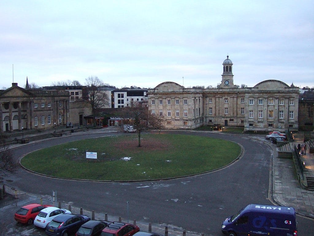 A circular green courtyard is bordered by historic buildings, parked cars, a tree in the center, and people walking nearby.