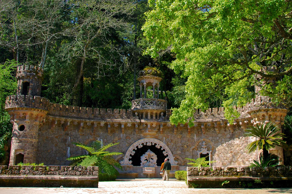 A person walks in front of a stone fortification surrounded by lush green trees and ferns, with a small pavilion on top.