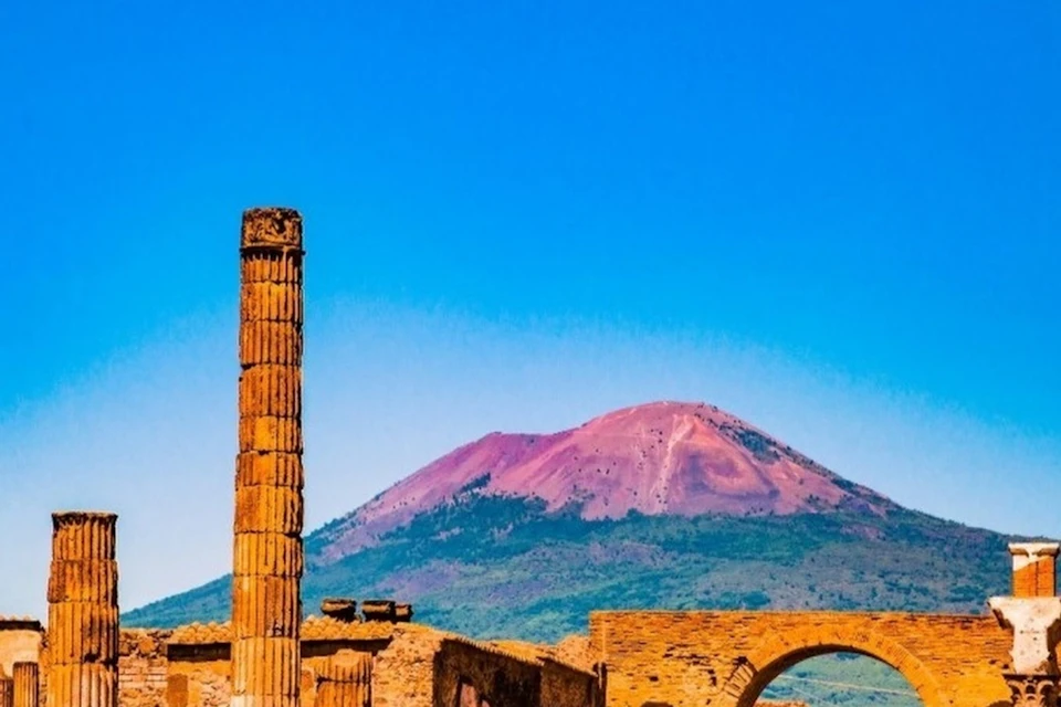 Ancient ruins with a column and an arch in the foreground, with a mountain under a clear blue sky in the background.