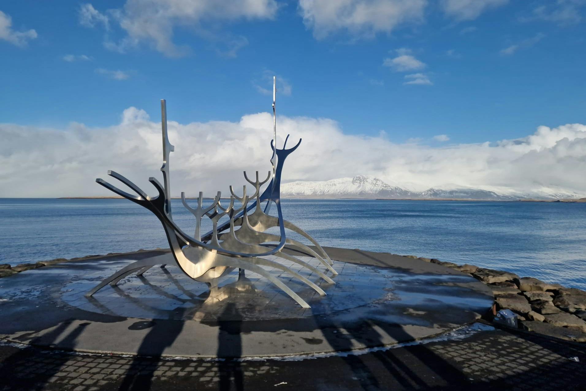 Metal sculpture resembling a boat with tall, curved structures, set against a backdrop of water and snow-capped mountains under a blue sky.