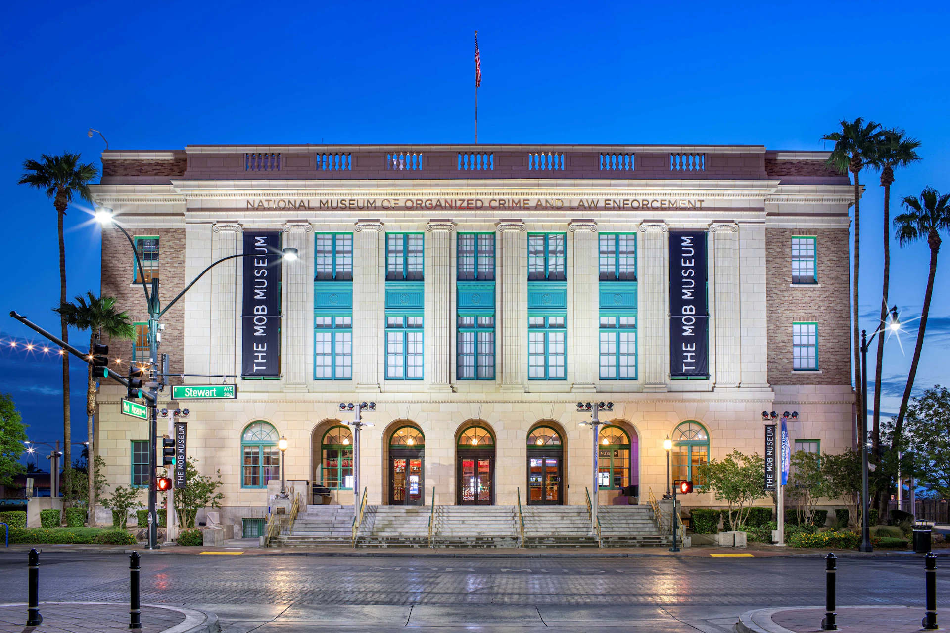 Exterior of the Mob Museum, a multi-story building with large arched windows, banners, and a neon sign. The sky is clear and blue.