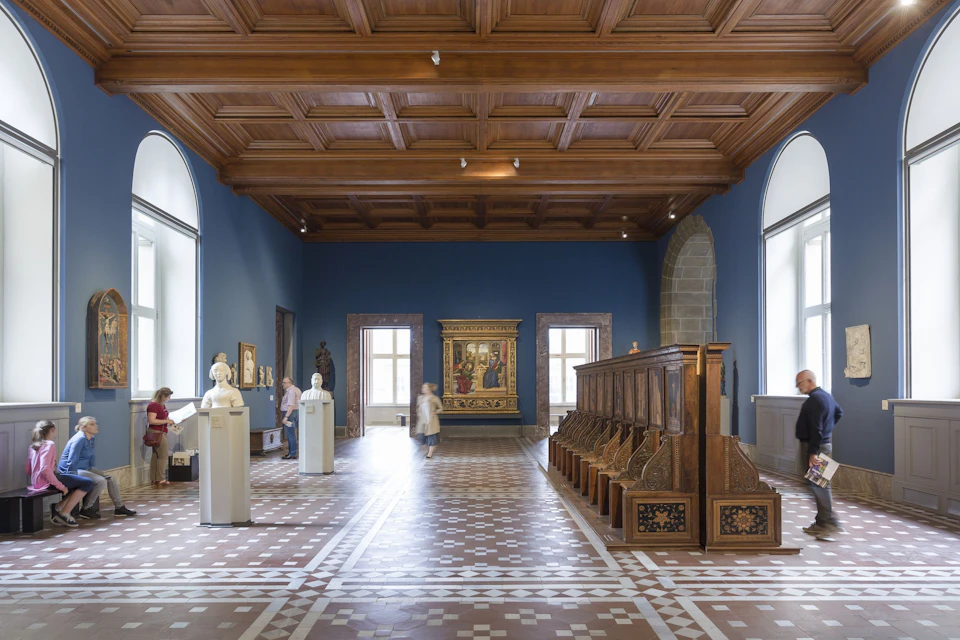 A museum room with wooden ceiling, statues on pedestals, a wooden choir stall, and a painting on a blue wall.