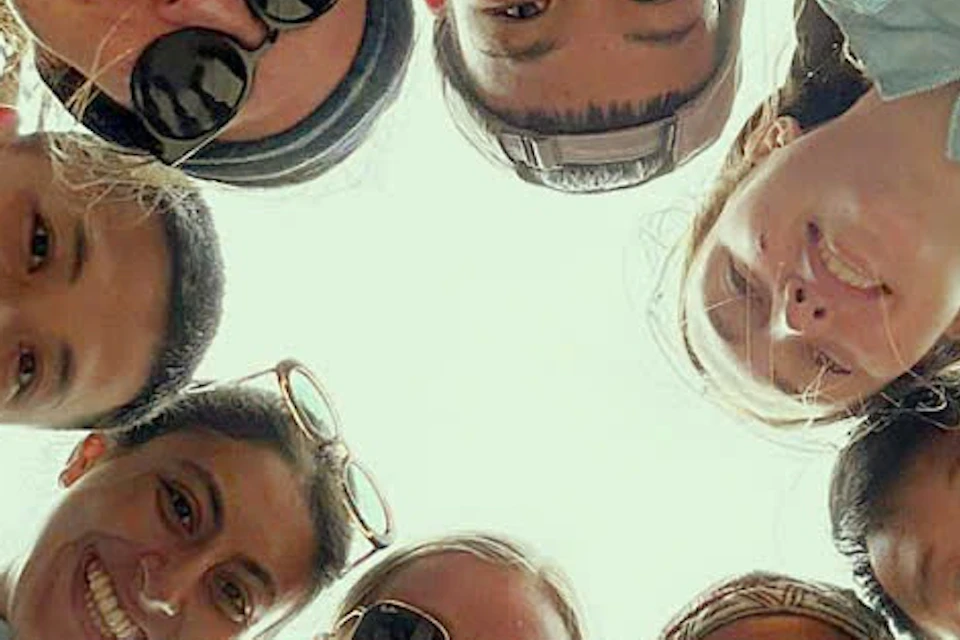A group of seven smiling people, some wearing sunglasses and blue jackets, huddled in a circle, looking down at the camera.