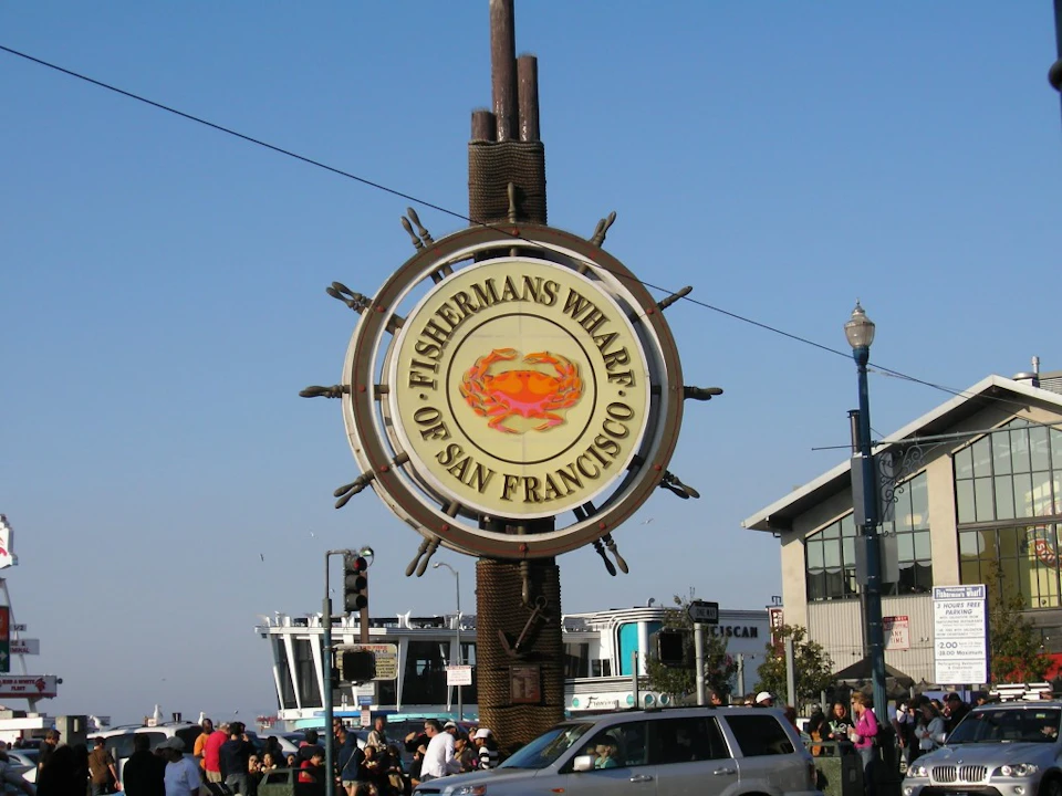 Large Fisherman's Wharf sign with crab logo, people, cars, and buildings in the background under a clear sky.