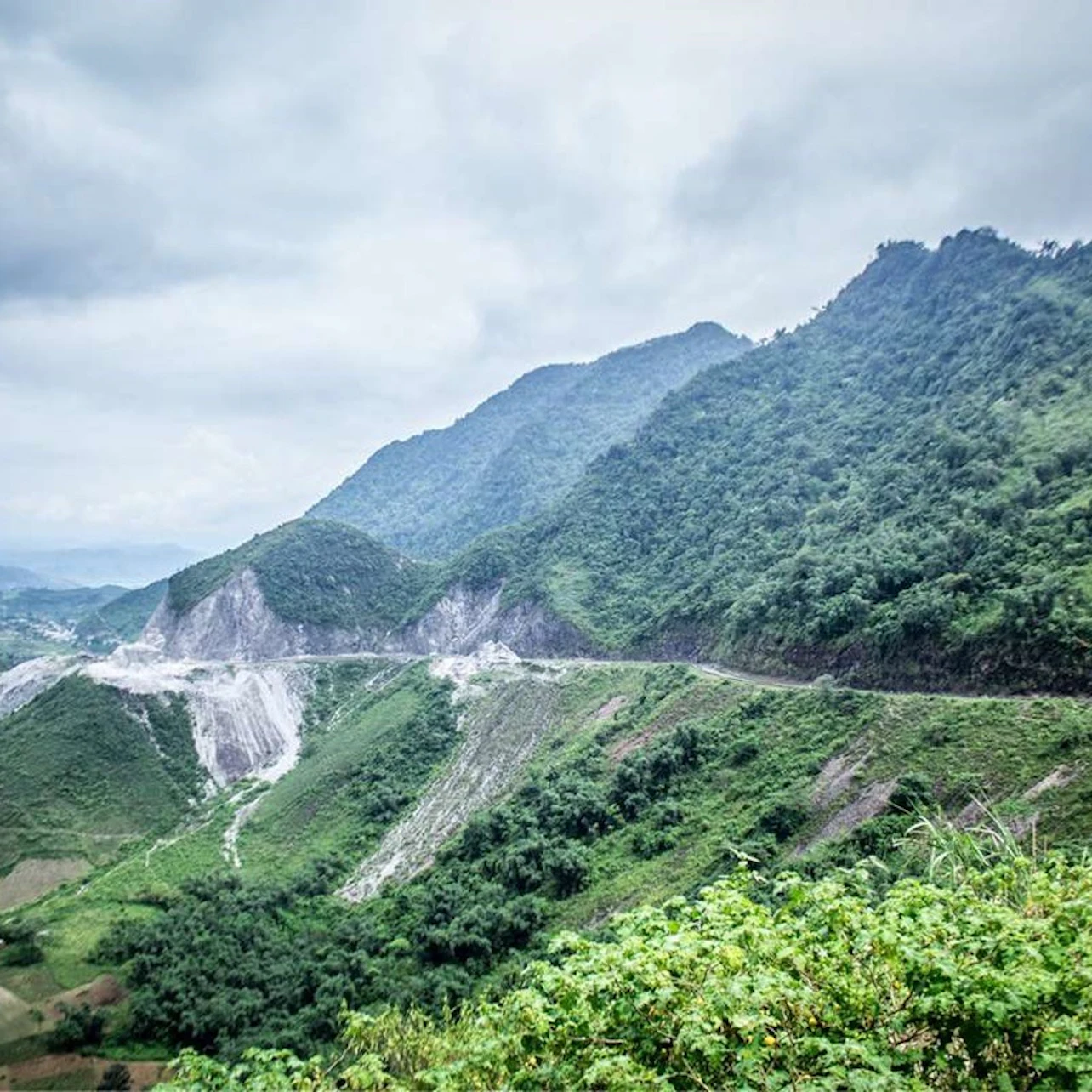 A winding road cuts through lush green mountains under a cloudy sky. The landscape features dense vegetation and hillside slopes.