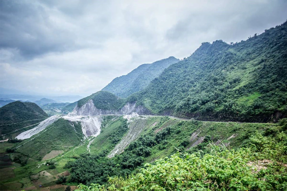 A winding road cuts through lush green mountains under a cloudy sky. The landscape features dense vegetation and hillside slopes.