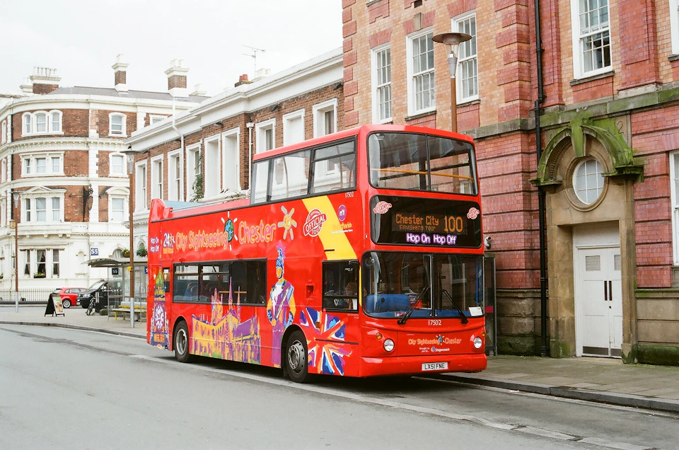 A red double-decker bus decorated with Chester city landmarks and Union Jack flags is parked on a street beside brick buildings.