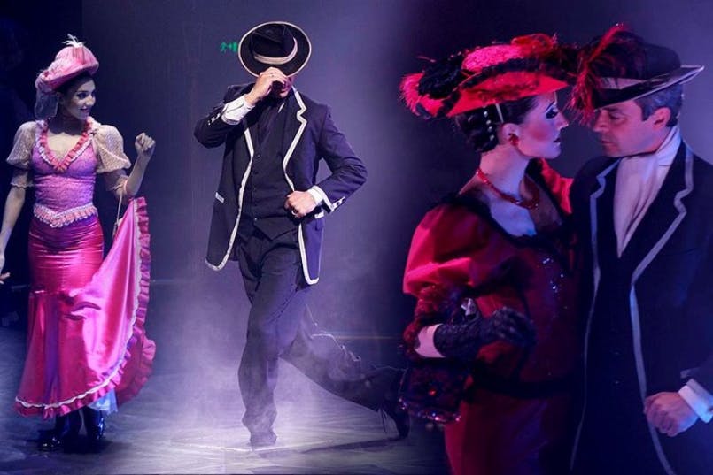 A man in a black suit and hat dances with a woman in a pink and purple dress, with another woman in red nearby on stage.