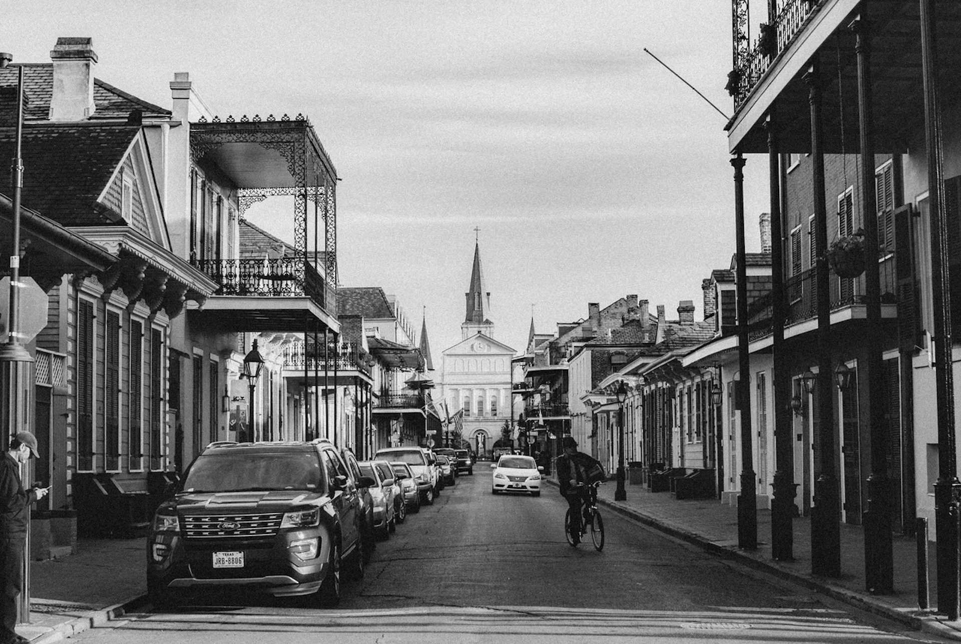 Black and white street view with parked cars, a cyclist, and buildings leading to a cathedral in the distance.