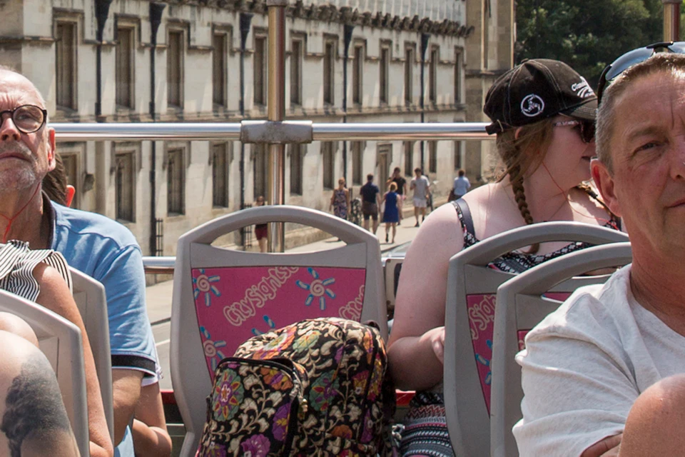 Tourists on a double-decker bus tour, with historic stone buildings and a tall church tower in the background under a clear sky.