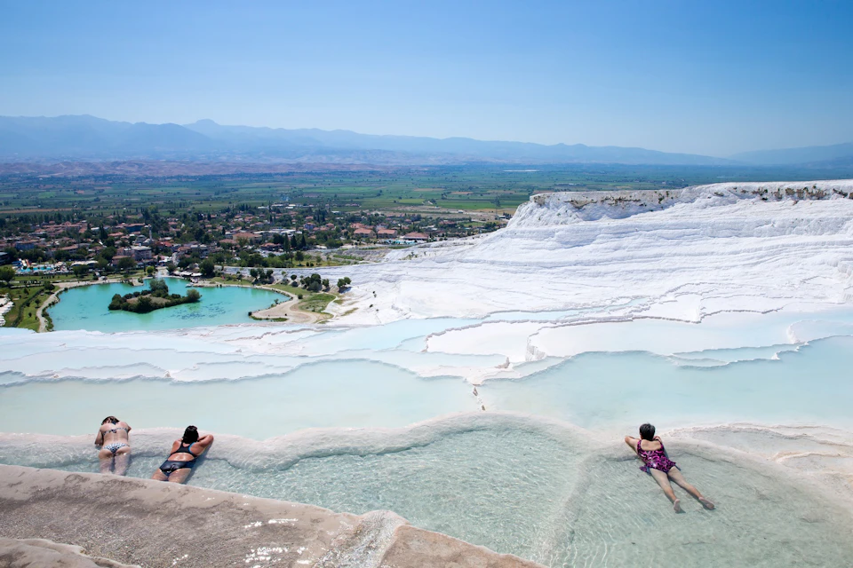 Two people relaxing at the edge of natural thermal pools with a scenic view of a town and mountains in the distance.