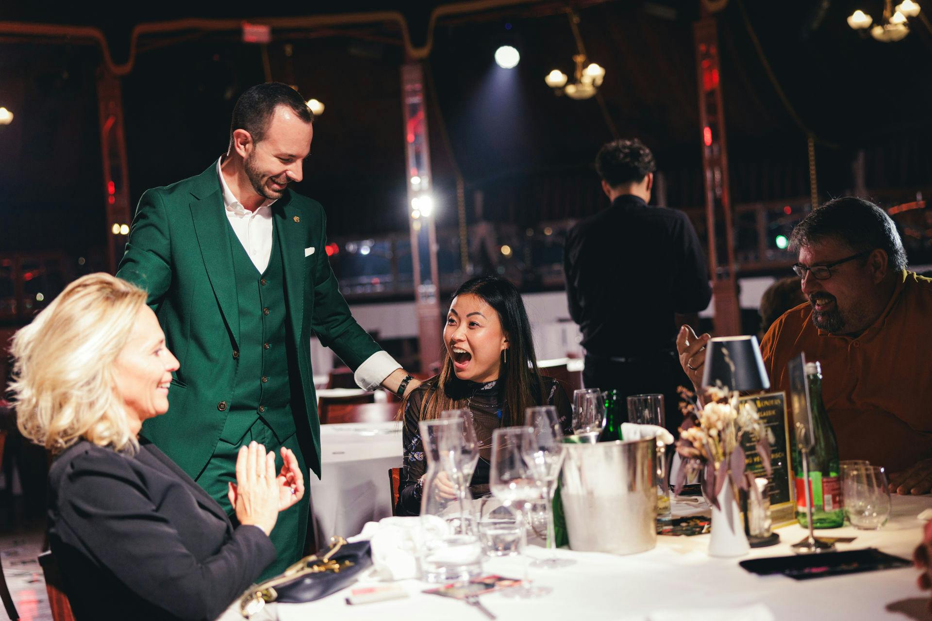 一位女士坐在餐桌旁，对着一位身穿绿色西装的男士惊讶地微笑；可以看到其他用餐者和空杯子。